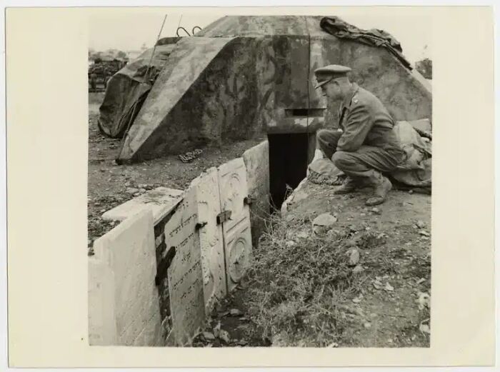 A historical photo of a man in uniform crouching by a bunker entrance, almost impossible to explain.