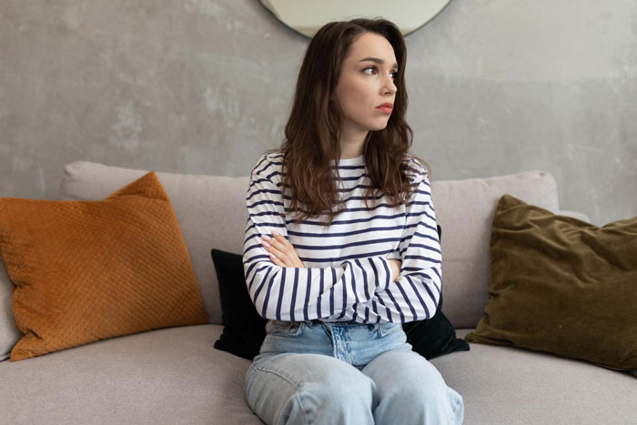 A young woman with long brown hair, wearing a striped shirt and jeans, sits on a couch with her arms crossed, looking upset. A young woman with long brown hair, wearing a striped shirt and jeans, sits on a couch with her arms crossed, looking upset.