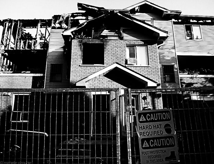 Black and white image of a burned creepy house behind a metal fence with caution signs