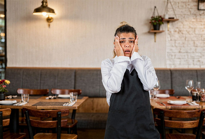 A scared waitress at a restaurant, hands on her face, after a lie about an allergy emergency.