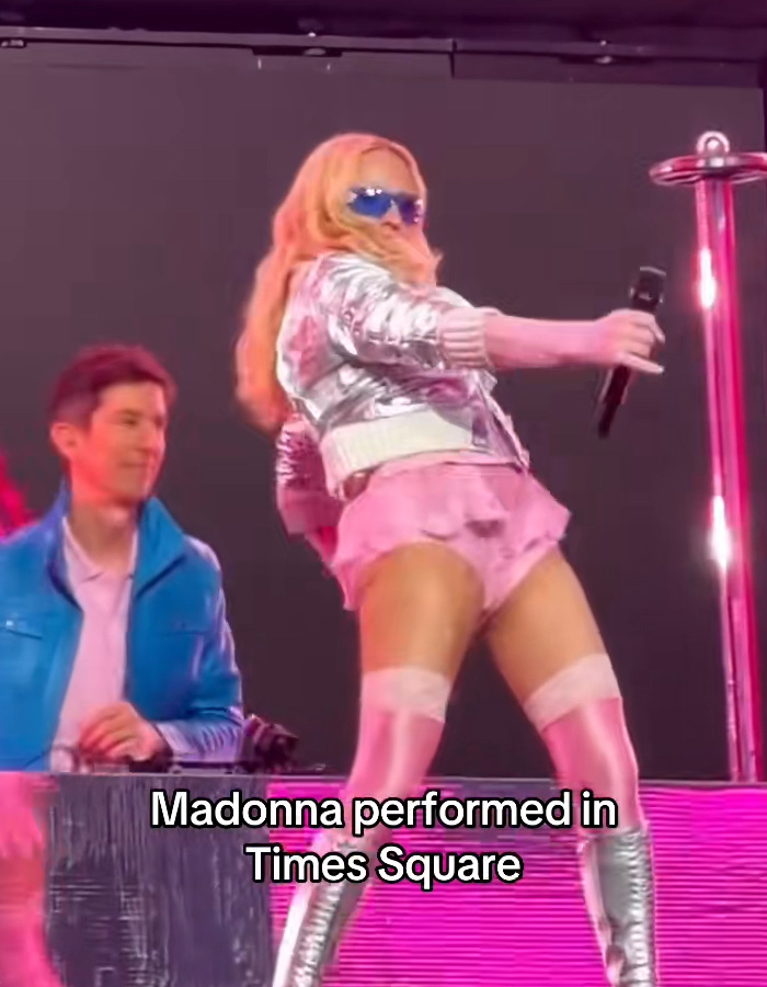 Madonna's Times Square performance. Madonna in a silver jacket and pink skirt, holding a microphone, with a person behind a DJ booth.