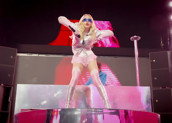 Madonna's Times Square performance. Madonna in a silver jacket, pink skirt, and boots performs on a brightly lit stage.