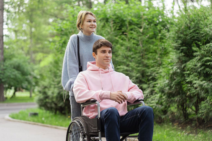 A woman pushes her ex-husband in a wheelchair, illustrating his accident and her continued support.