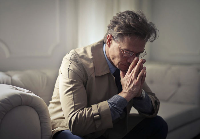 A man with glasses in a beige trench coat, looking down with hands clasped, expressing sadness about his marriage ending.
