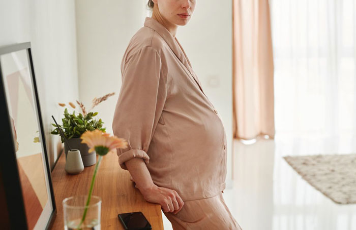A pregnant woman in neutral pajamas stands near a dresser, reflecting on her husband's accident and marriage.