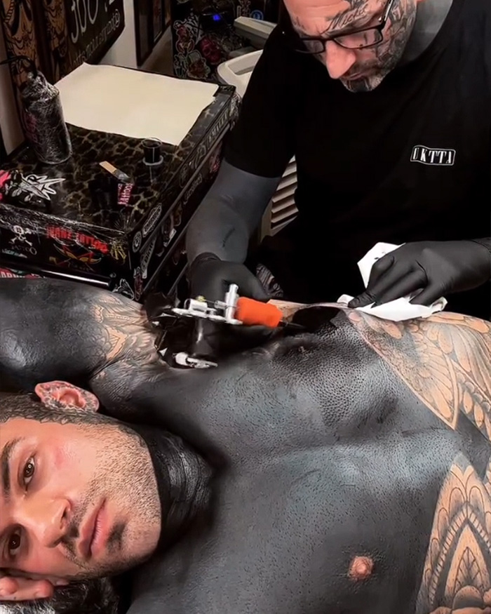 Man getting blackout tattoos on his chest and neck, lying on a tattoo artist's table while the artist works.