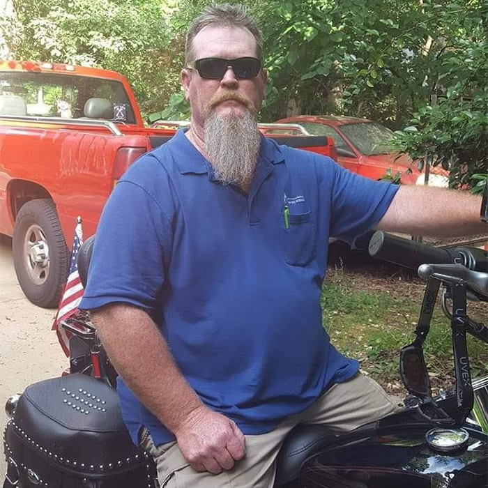 Man loses his temper, sitting on a motorcycle, wearing sunglasses and a blue shirt.