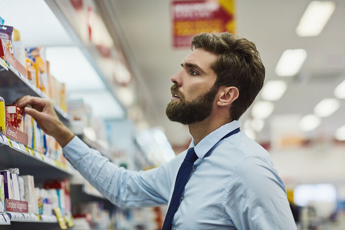 A man with a beard in a blue shirt and tie, thoughtfully reaching for an item on a shelf, showing support for a partner.