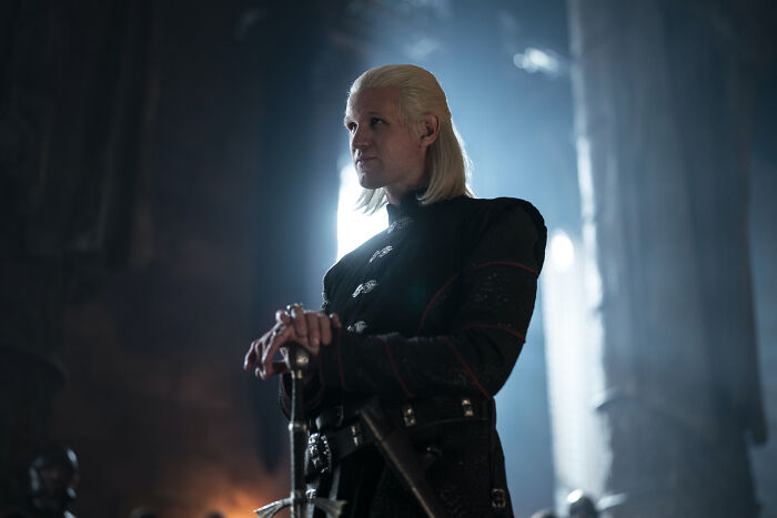 A man with white hair wearing black attire holds a sword in a dark hall in House Of The Dragon, a disturbing moment.