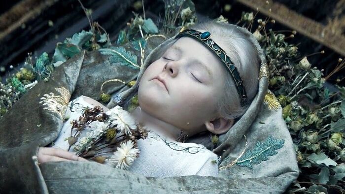 A young child with white hair and a crown lies peacefully in a bed of flowers in House Of The Dragon, a disturbing moment.