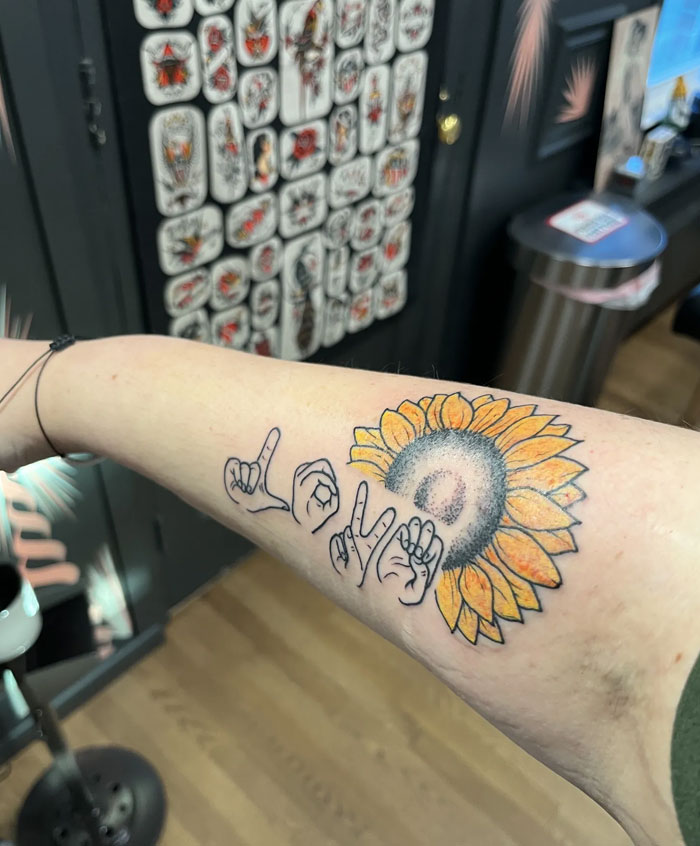 A colorful sunflower tattoo intertwined with the word LOVE spelled out in sign language, an artistic and meaningful design.