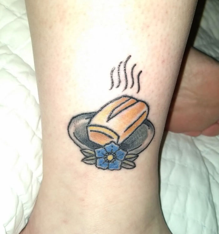 A unique tattoo on an ankle featuring a hot dog in a bun with a blue flower.