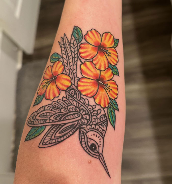 A colorful hummingbird tattoo with vibrant orange hibiscus flowers on an arm.
