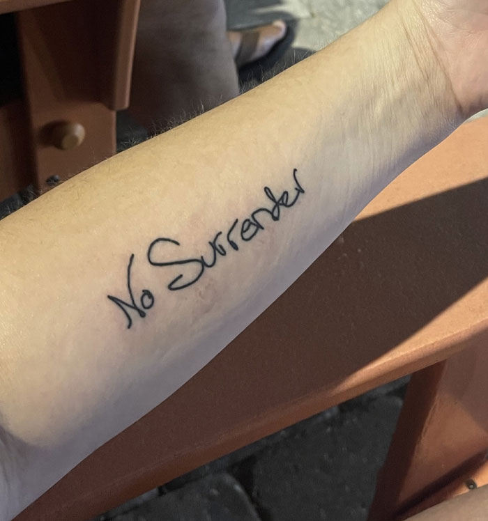 An arm with a significant tattoo of the phrase No Surrender in script handwriting.