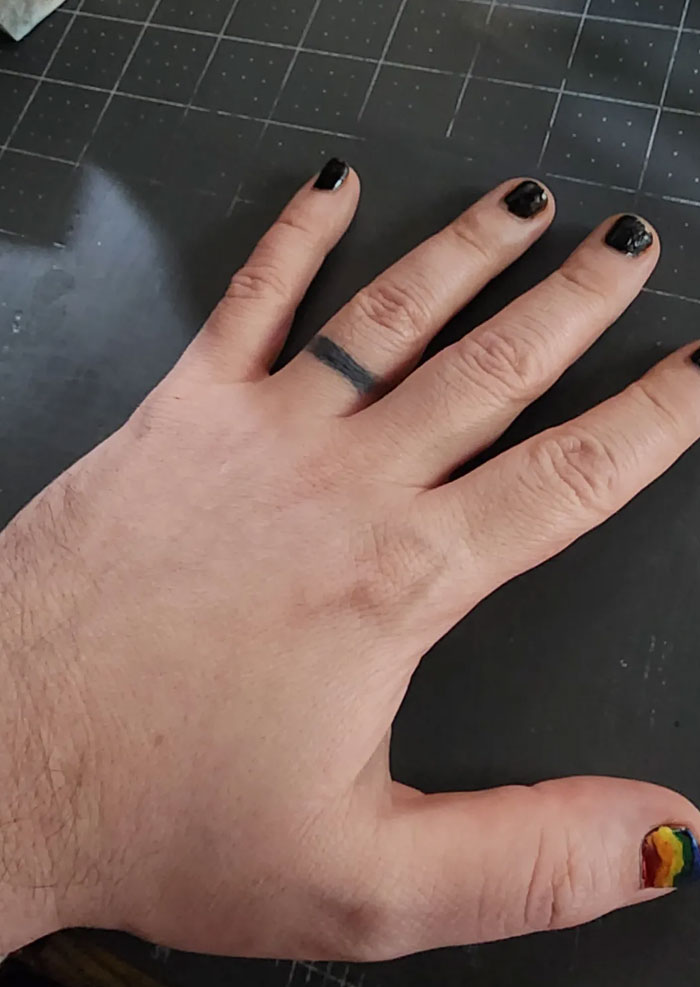A hand with dark painted nails and a simple black band tattoo on the ring finger. This tattoo means far more than what meets the eye, symbolizing commitment or memory.