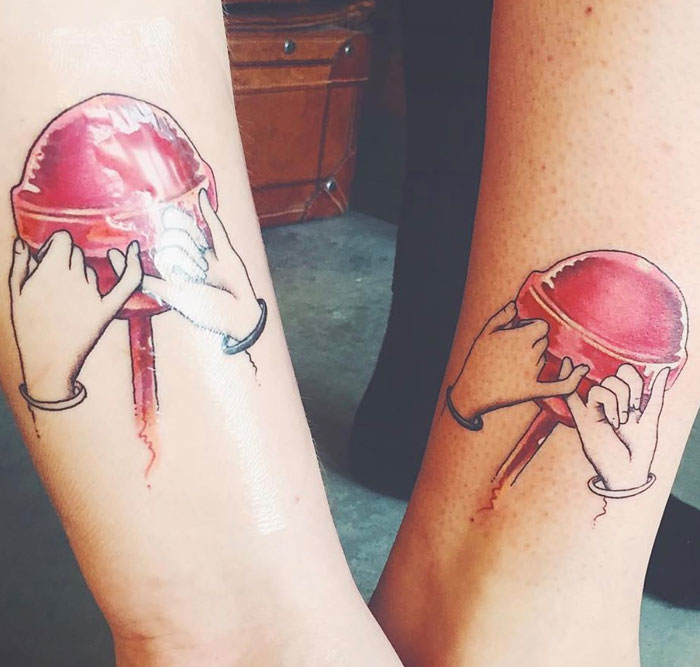 Two legs with matching tattoos of hands holding a red lollipop. These meaningful tattoos often symbolize friendship or connection.