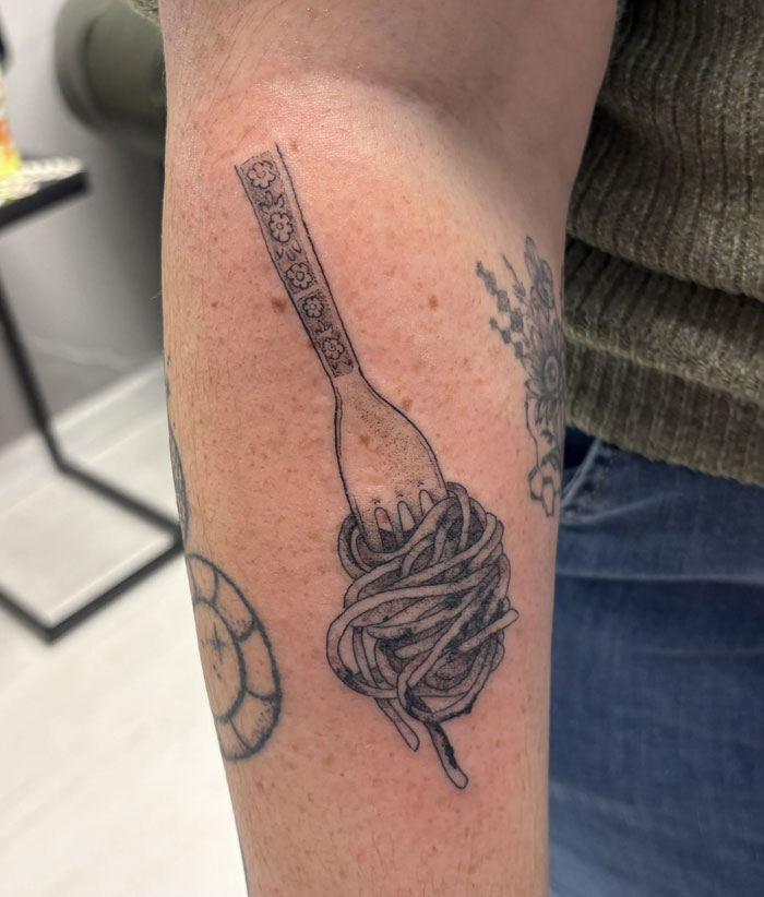 An arm featuring a detailed tattoo of spaghetti wrapped around a fork. This creative tattoo carries significant meaning.