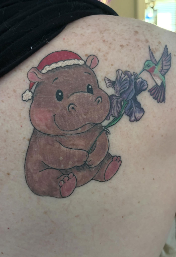 A charming hippo tattoo wearing a Santa hat, holding a flower, with a hummingbird nearby, showcasing meaningful tattoos.