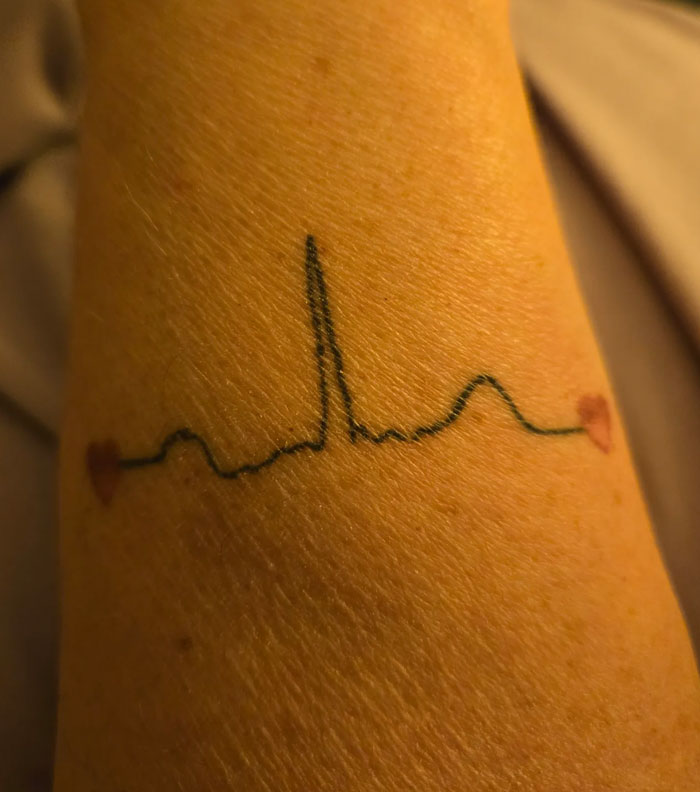 A close-up of a forearm reveals a minimalist heartbeat line tattoo, subtly adorned with two small hearts.