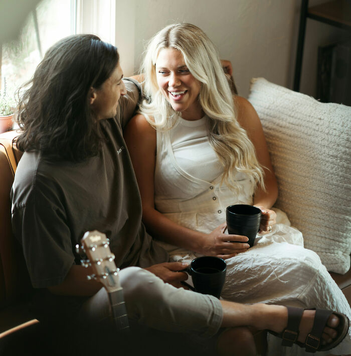 A couple sits on a couch, smiling and holding mugs, sharing an intimate moment to marry their partner.