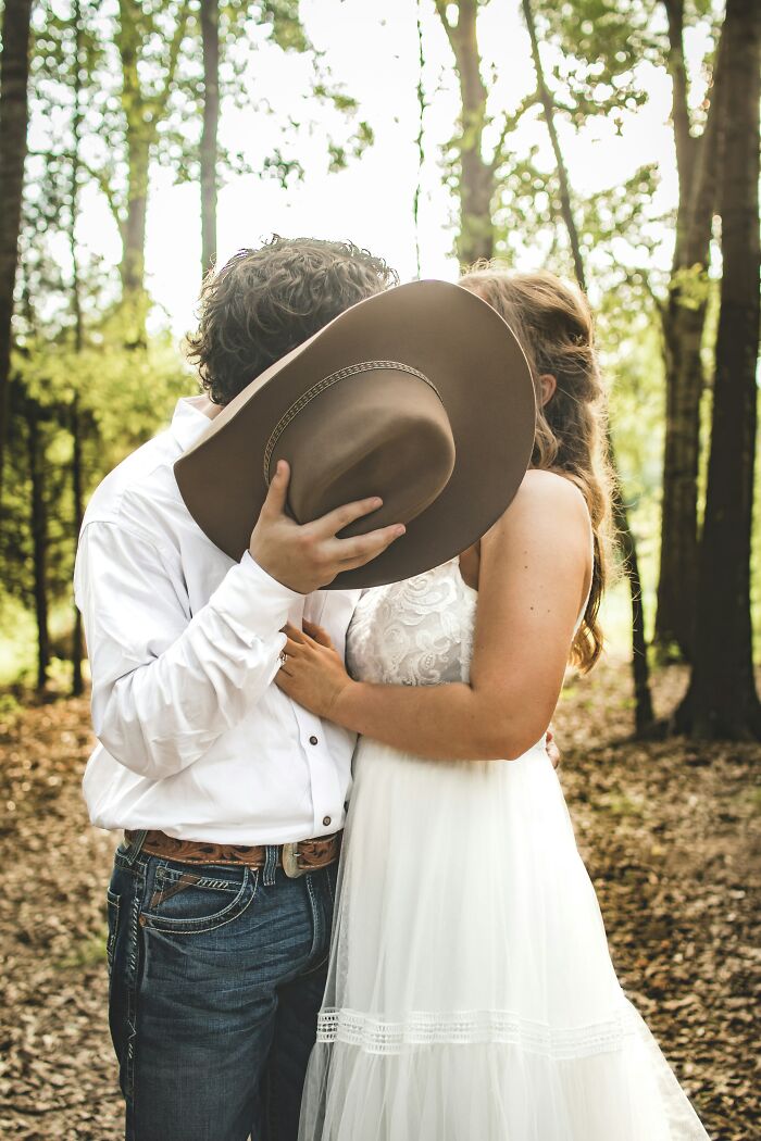 Couple in wedding attire hiding faces behind cowboy hat in forest