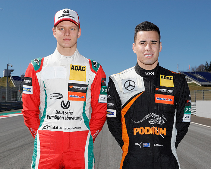 Two race car drivers standing on a track in racing suits with ADAC and Deutsche Vermögensberatung logos Two race car drivers standing on a track in racing suits with ADAC and Deutsche Vermögensberatung logos