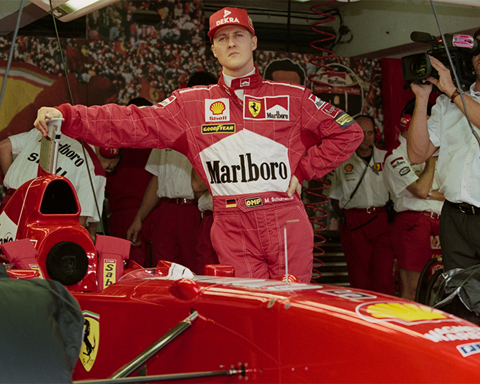 Michael Schumacher in red racing suit standing by Ferrari Formula 1 car in garage Michael Schumacher in red racing suit standing by Ferrari Formula 1 car in garage