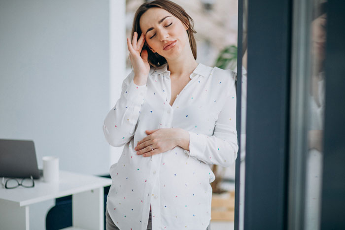 A pregnant DIL looking stressed, holding her head and baby bump, frustrated by her MIL's demands on the baby's name.