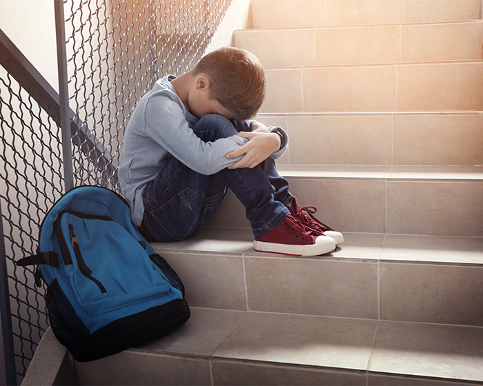 A boy sits on stairs, hunched with a backpack beside him, showing changed behavior, after mom hid audio recorder.