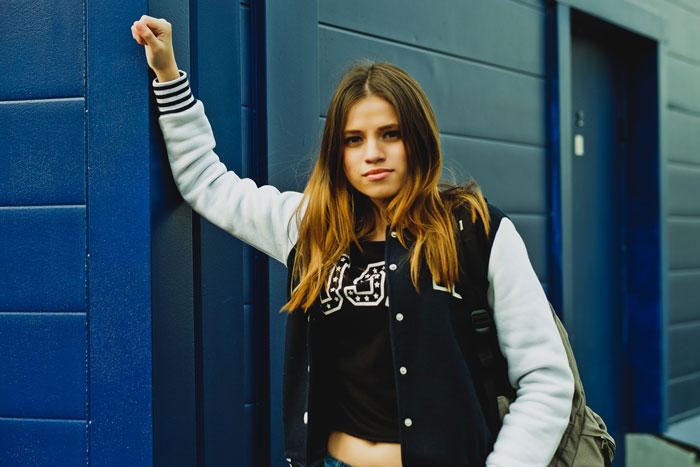A teen girl in a black and white letterman jacket with a backpack, leaning against a blue wall, related to a religious mom.