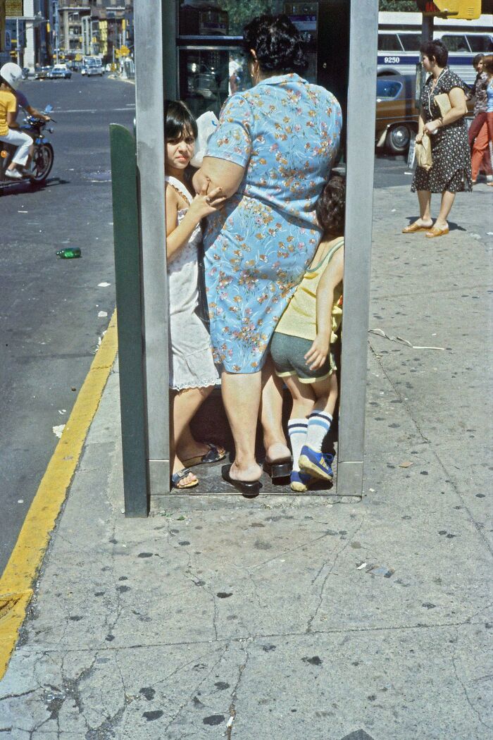 Candid street photo of a crowded phone booth with woman and children in vintage clothing