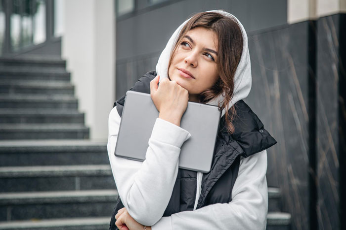 A young woman, 18YO, with a thoughtful expression, holding a laptop, considering her parents' hostile expectations. A young woman, 18YO, with a thoughtful expression, holding a laptop, considering her parents' hostile expectations.