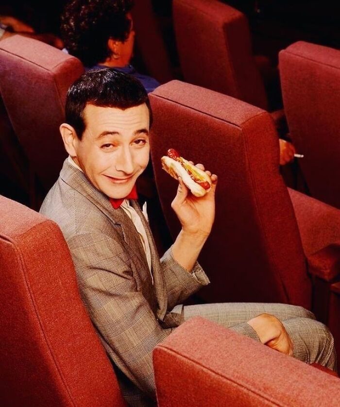 Pee-wee Herman in a suit holding a hotdog, a memorable moment among celebrity scandals.