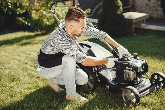 A man in a checked shirt and an apron, tending to a lawnmower, not a joke for people.