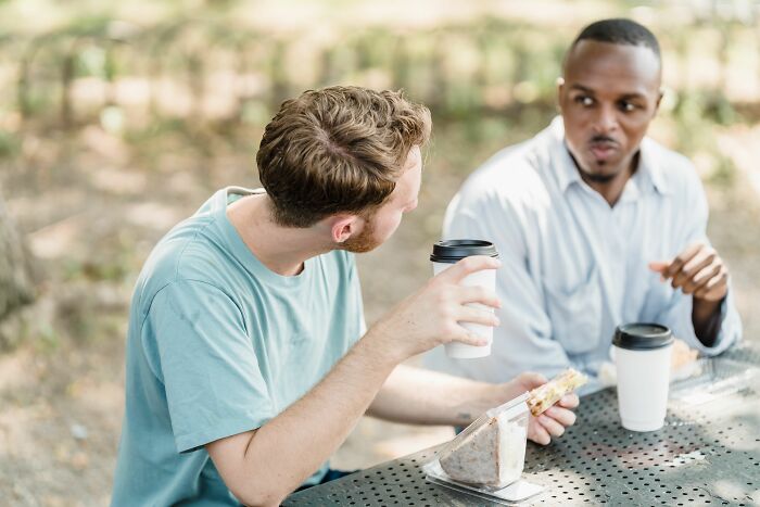 Two men sitting at a table outdoors, one looking surprised, realizing something was not a joke.
