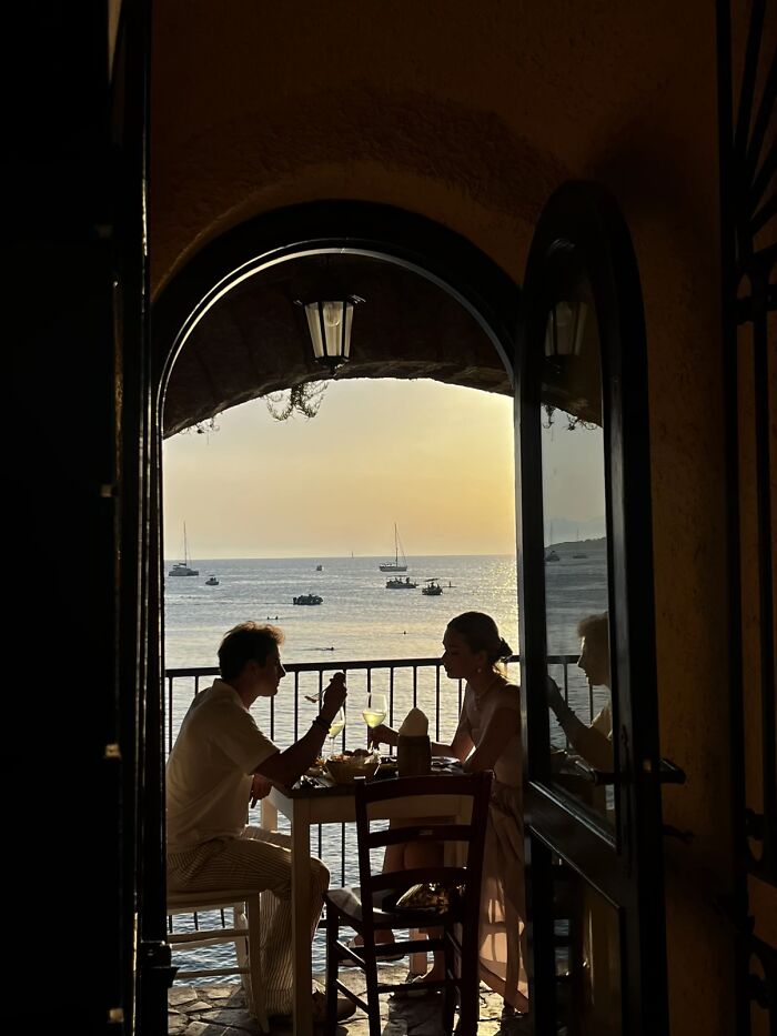 A couple dining on a balcony overlooking the sea at sunset, a serene scene from the wholesome photo trend.