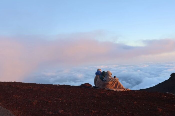 Two people bundled in a blanket sit on a mountain peak, looking at clouds, embodying the wholesome photo trend.