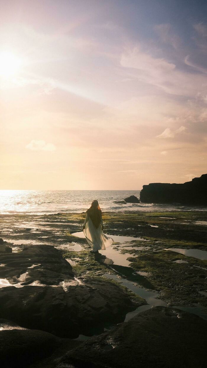A person in a flowing dress walks on a rocky beach at sunset, an example of the wholesome photo trend.