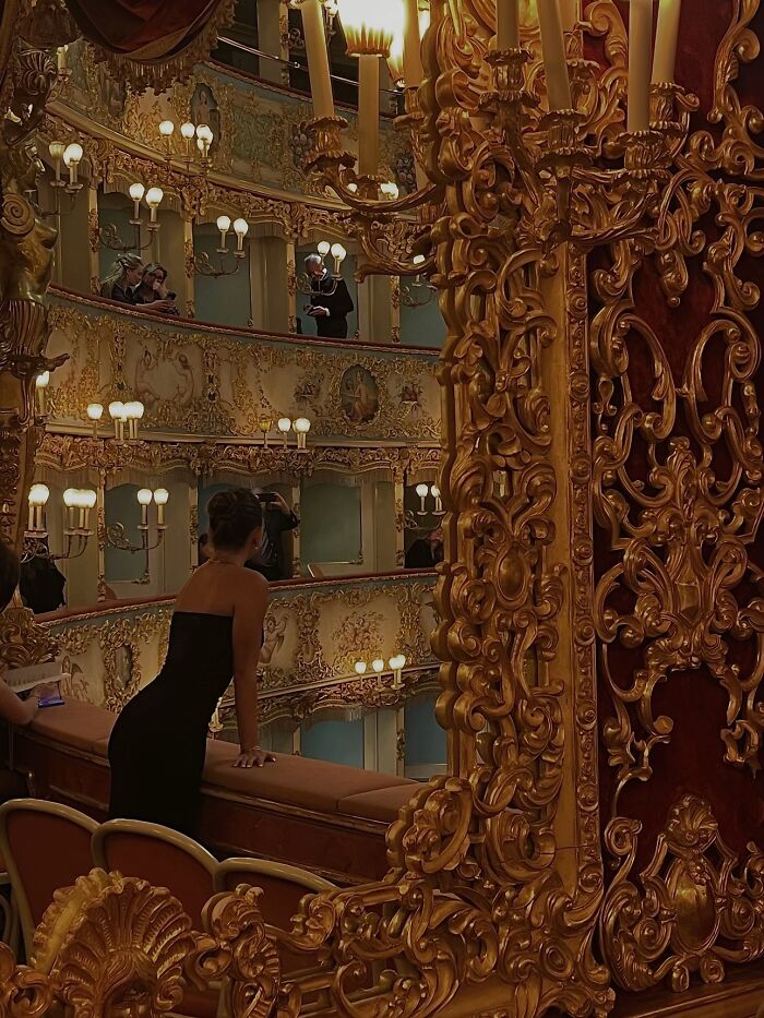 A woman in a black dress looks over a lavish opera house balcony, capturing the wholesome photo trend.