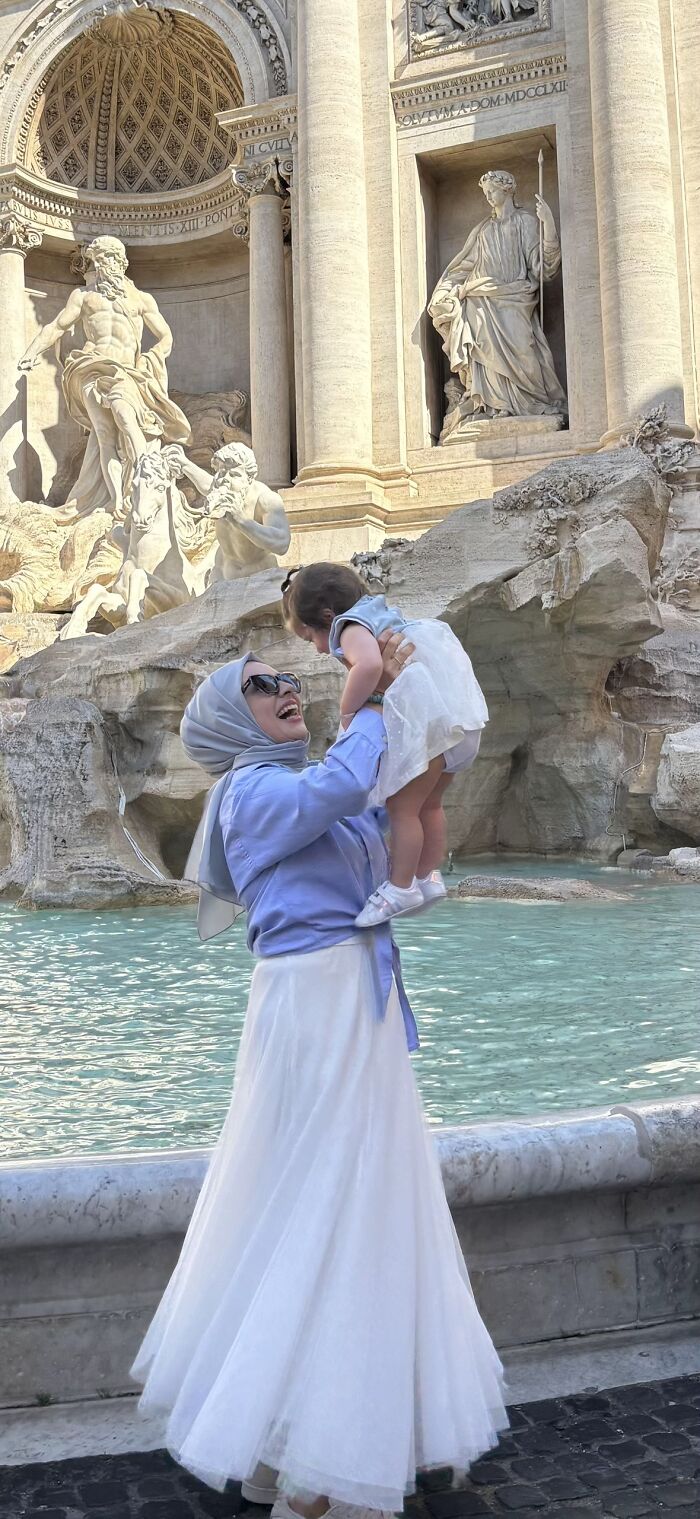 A mother in a hijab and white skirt lifts her baby joyfully in front of a fountain, a wholesome photo trend.