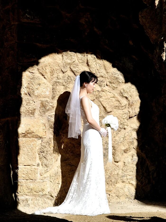 A woman in a white wedding dress stands under an arched stone wall, resembling a painting with light and shadow.