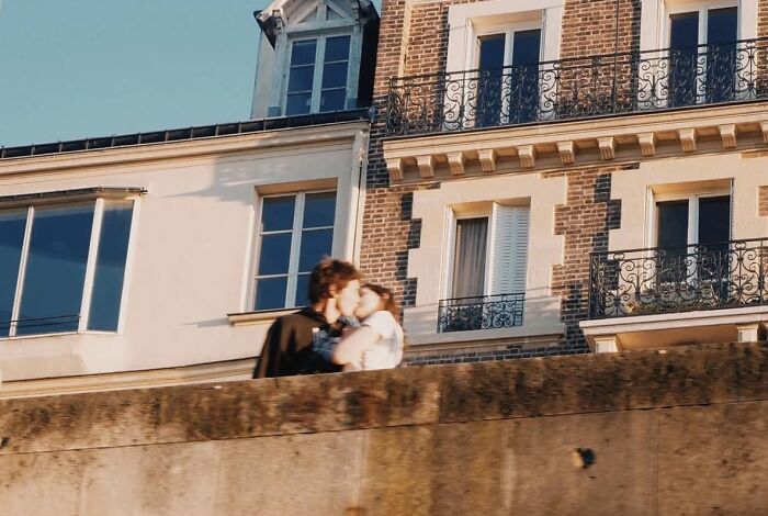 A couple kisses on a wall in front of charming European buildings, reflecting the wholesome photo trend.