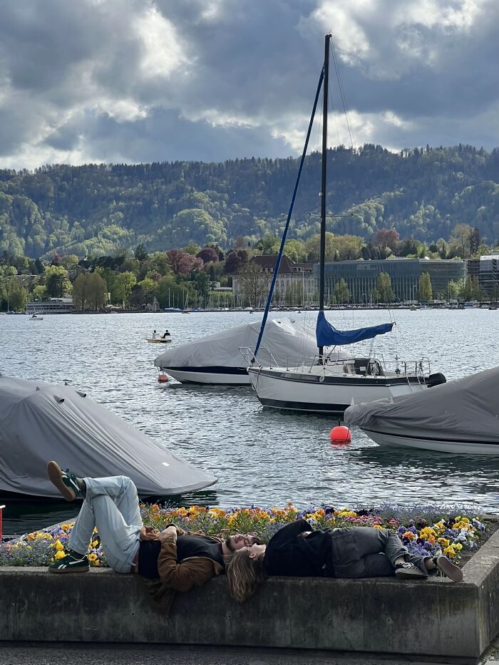 Two people lie by a lake with boats, surrounded by flowers, evoking the serene aesthetic of a painting photo trend.