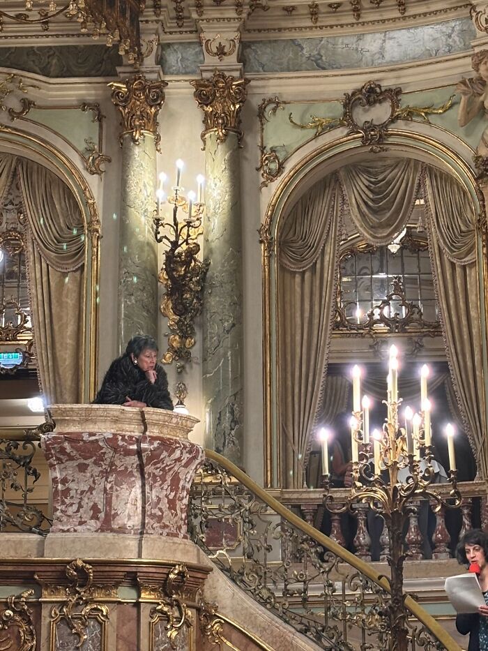 An older woman in a grand, ornate room looks down from a balcony, fitting the wholesome photo trend.