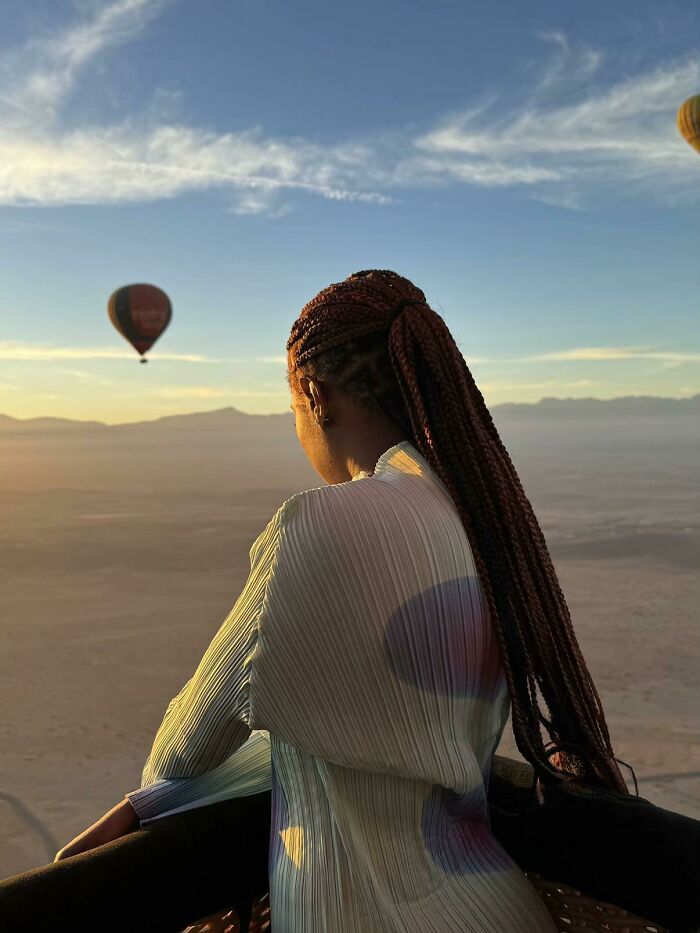 A woman with long braids looks out over a desert landscape with hot air balloons, embodying the wholesome photo trend.