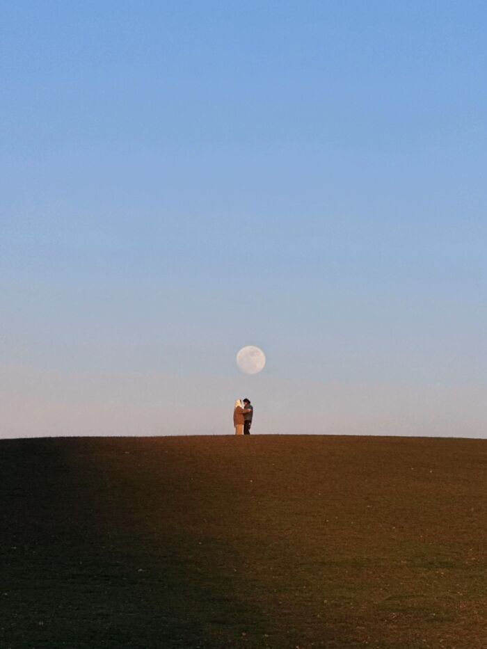 A couple stands on a hill, silhouetted against a large full moon, representing a wholesome photo trend.