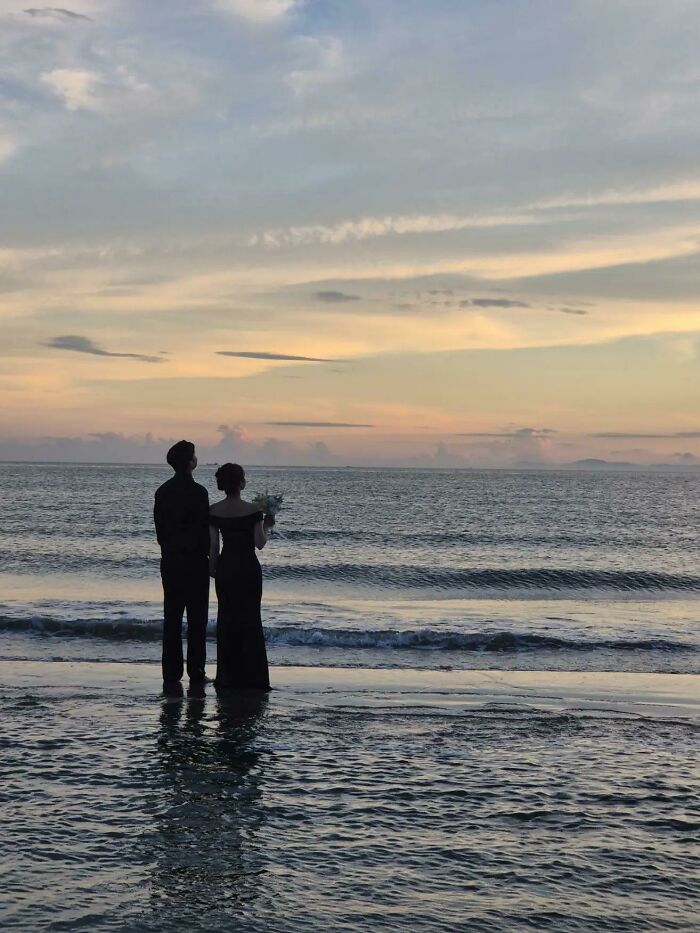 A couple in formal attire stands at the edge of the ocean, looking out at the sunset, embodying a wholesome photo trend.