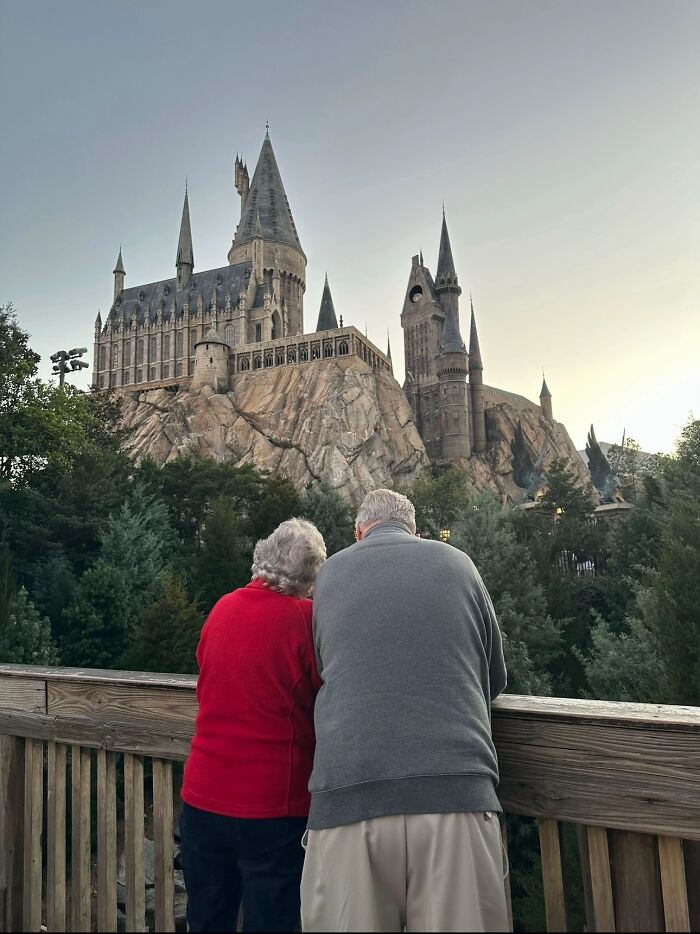 An elderly couple stands with their backs to the camera, looking at the Hogwarts castle, captured in a wholesome photo trend.