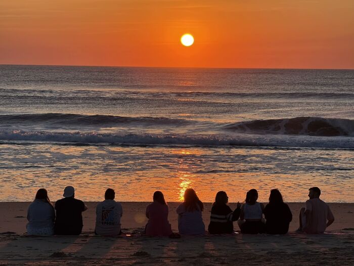 People watching a beautiful sunset at the beach, capturing the wholesome photo trend.
