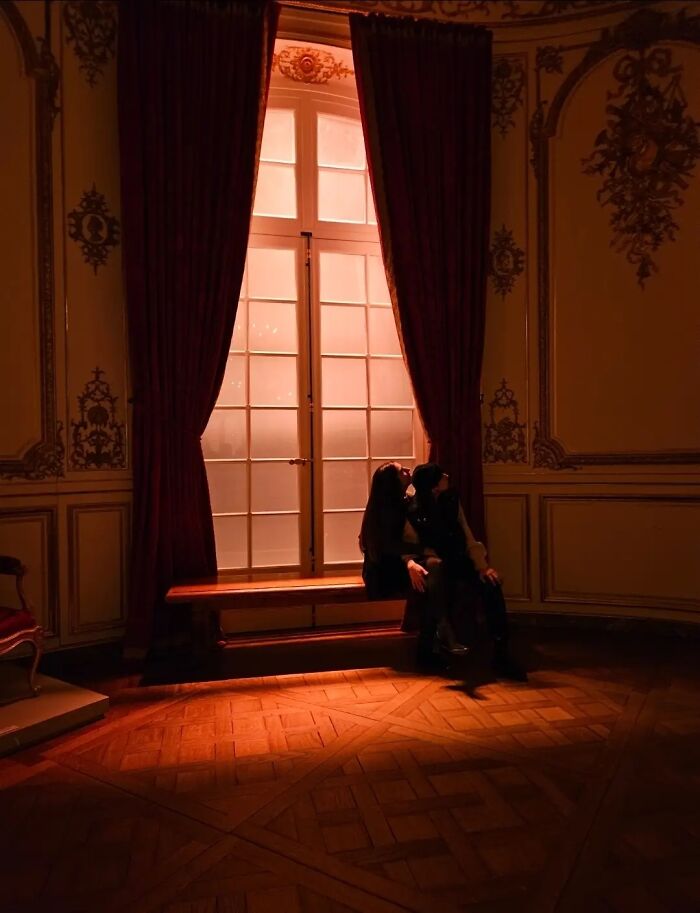 Two strangers sit by a window with red curtains, bathed in warm light, exemplifying the wholesome photo trend.